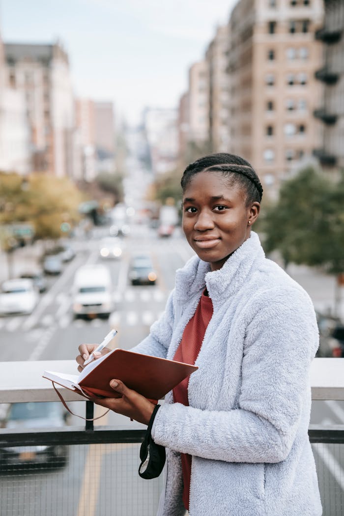Delighted young African American female student looking at camera while standing on footbridge over road and taking notes in notepad