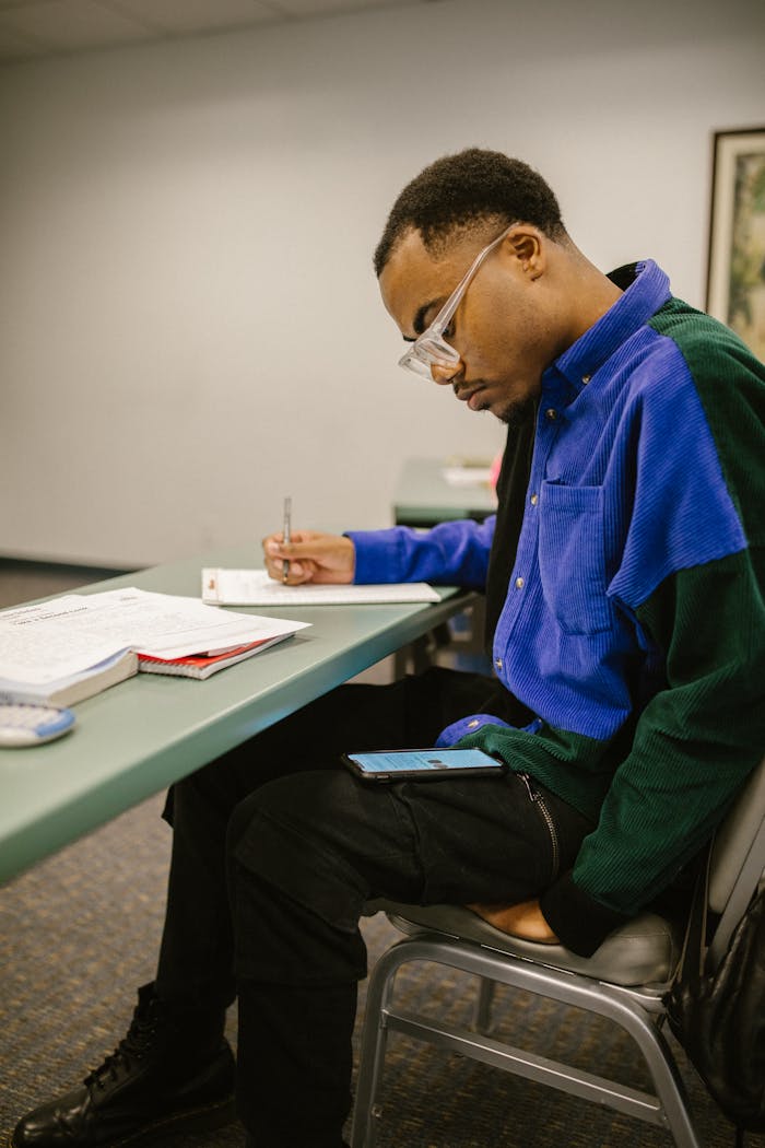 Black male college student writing in class with a smartphone on his lap, possibly cheating.